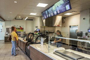 A fast-food restaurant counter with a staff member serving a customer, and menu screens overhead.