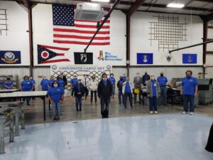 People are gathered in a facility with various flags and a large U.S. flag. They're posing and wearing matching shirts.