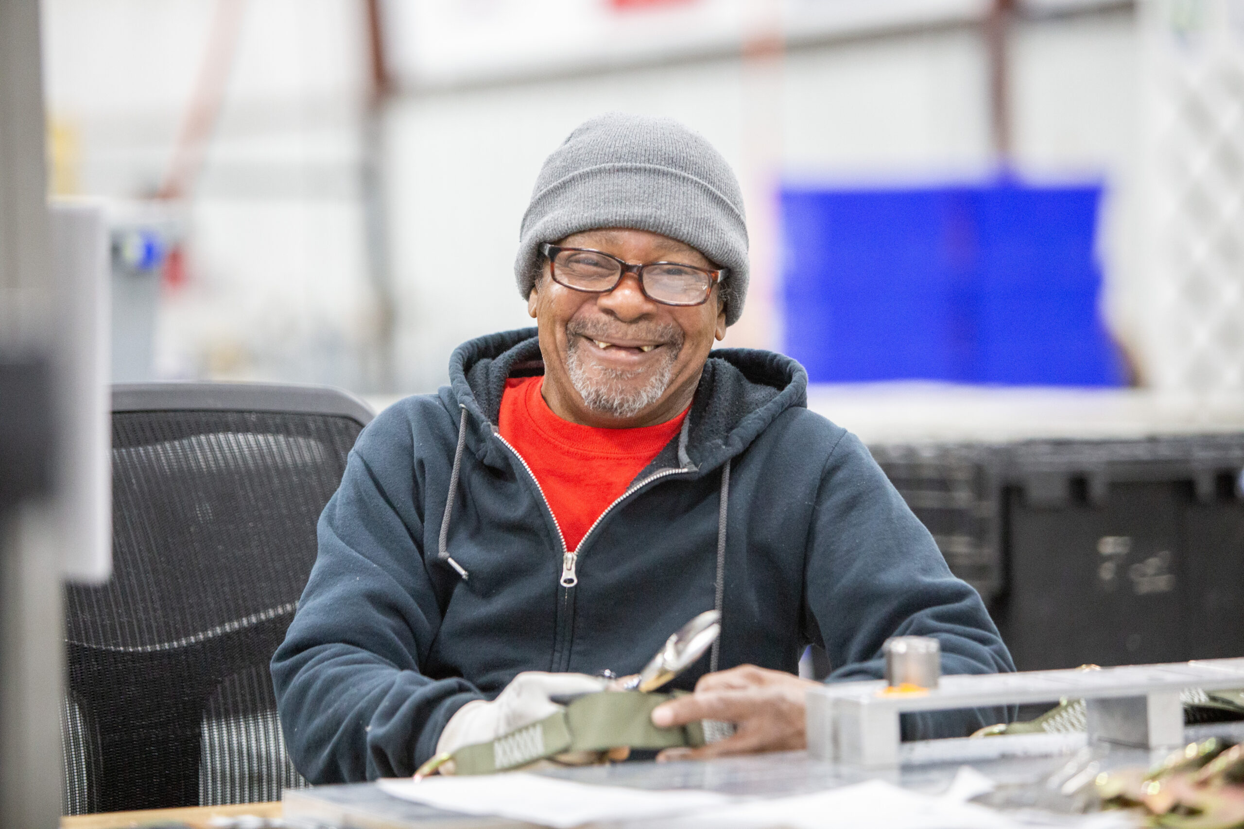 A person wearing a beanie and glasses is smiling while sitting in a workspace.