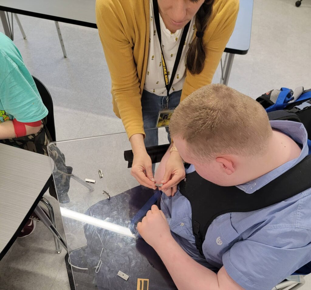 A person is assisting a seated individual with a task involving small objects on a table.
