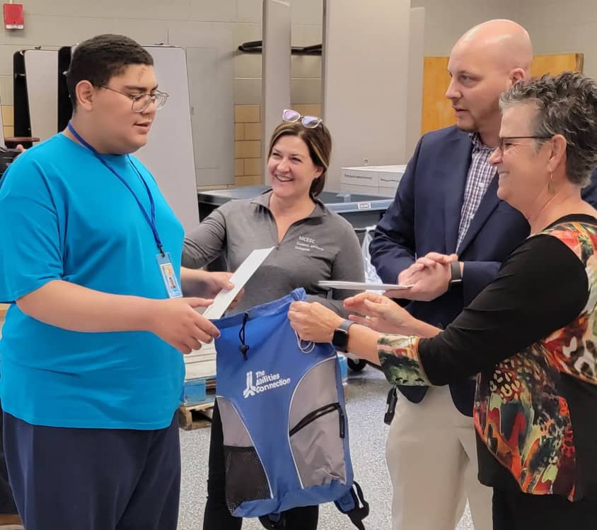 A group of people are smiling and holding papers and a bag labeled "The Abilities Connection."