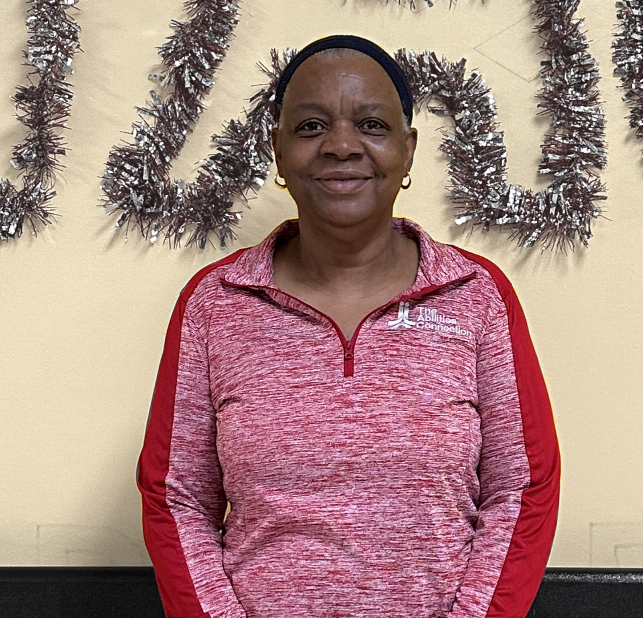 A person wearing a red pullover stands in front of a wall with tinsel decorations.