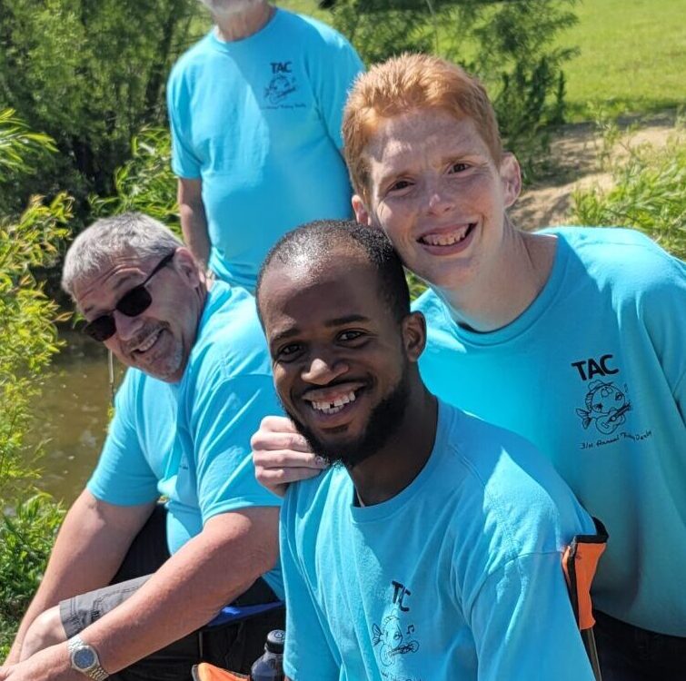 A group of people smiling and wearing matching blue T-shirts, outdoors by a stream or river.
