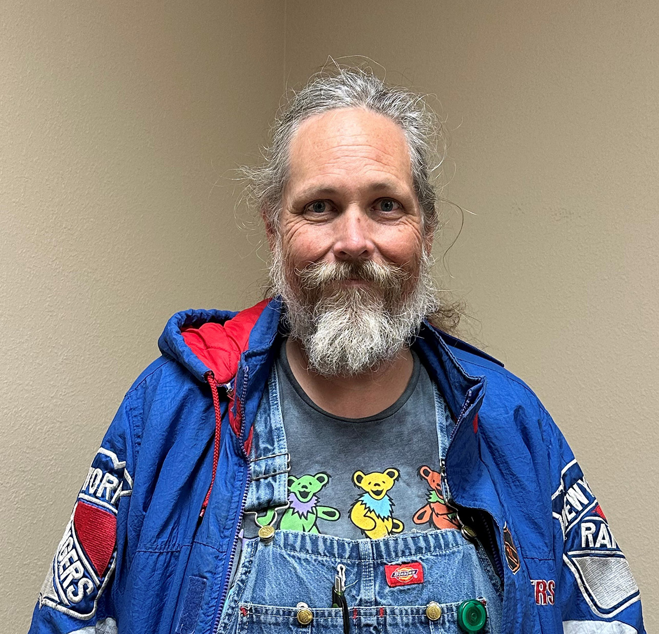 A person with a beard, wearing a colorful jacket and patterned shirt, smiles at the camera.