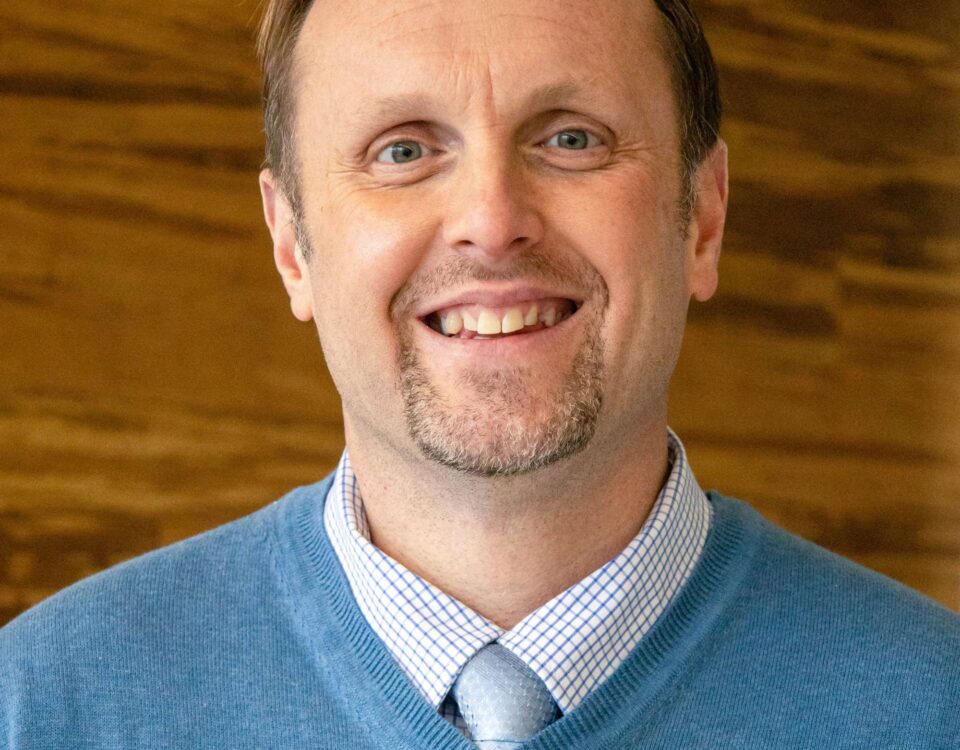 A person wearing a blue sweater, light blue shirt, and tie, smiling in front of a wooden background.