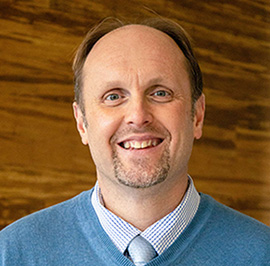 A person wearing a blue sweater, light blue shirt, and tie, smiling in front of a wooden background.
