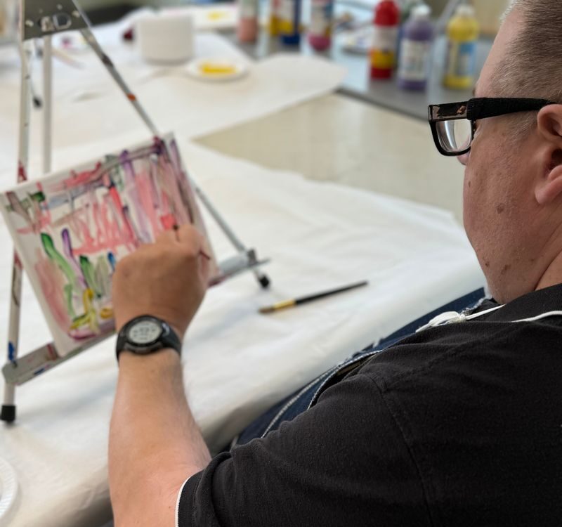 A person is painting on a small canvas using an easel, surrounded by art supplies.