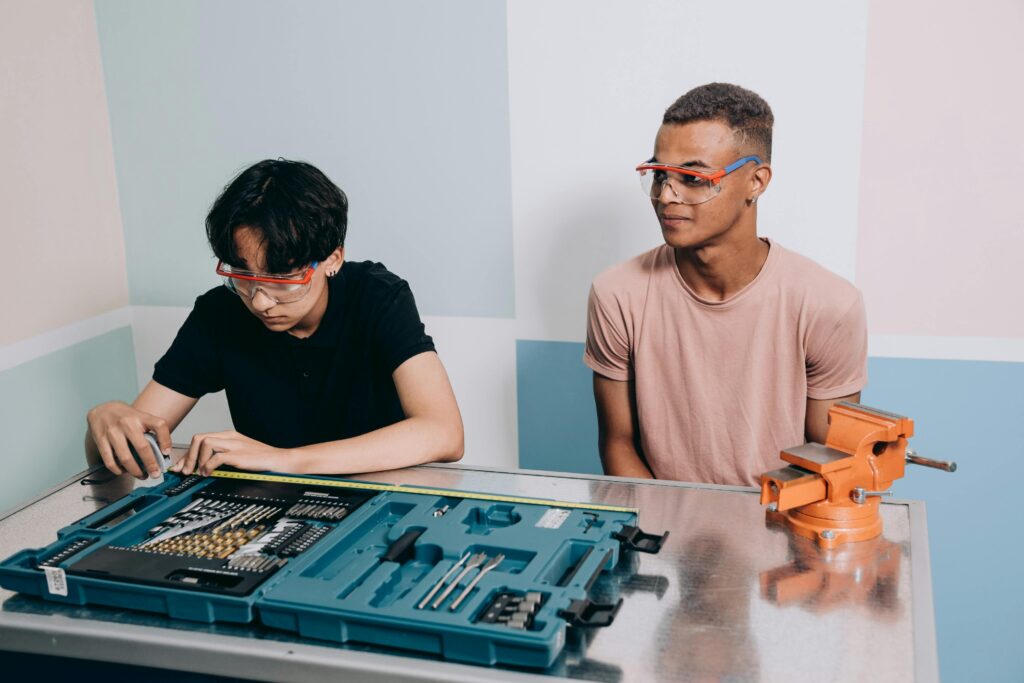 Two people wearing safety glasses are at a table with a toolkit and a vise, working on a project.