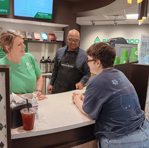 Three people are at a coffee counter, engaging in conversation.