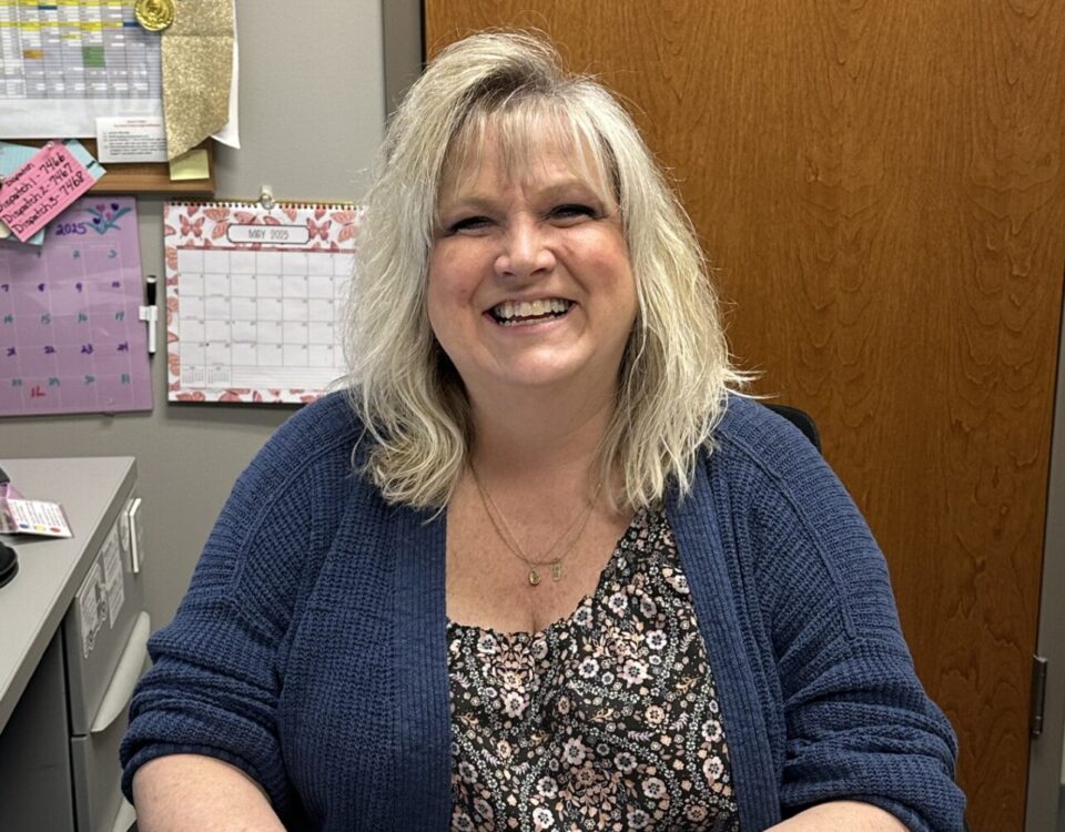 A person smiling while seated at a desk with calendars in the background.