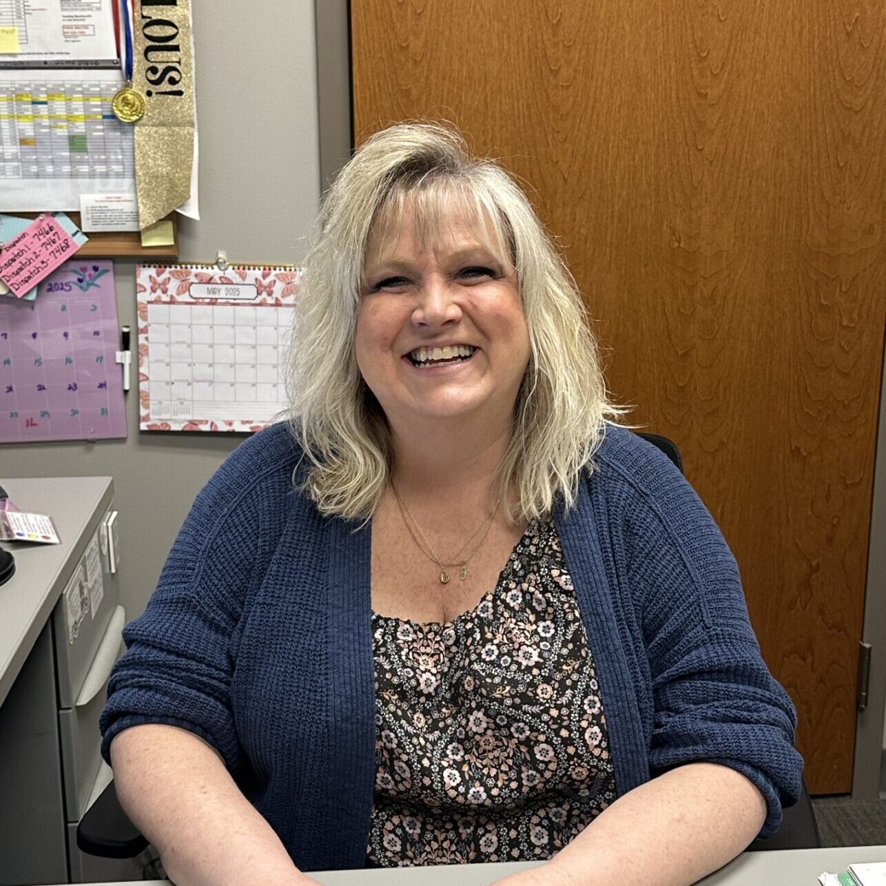 A person smiling while seated at a desk with calendars in the background.