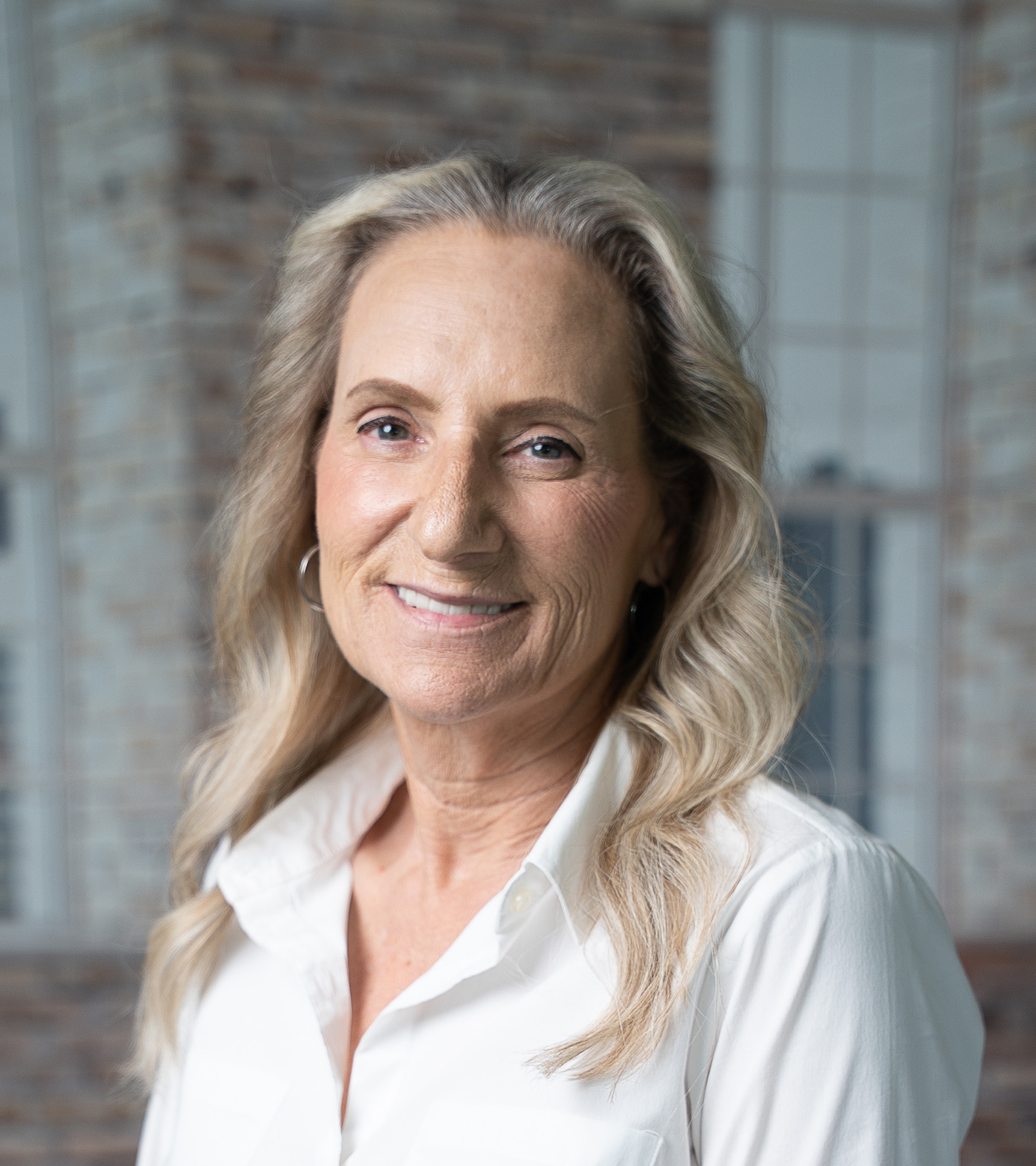 A person with long, blonde hair and a white shirt is smiling in front of a brick wall background.
