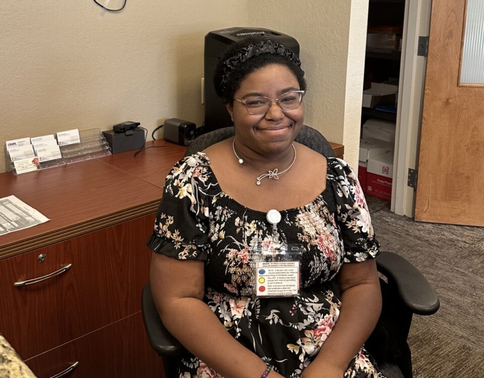 A person is sitting in an office setting, wearing a floral dress and a badge.