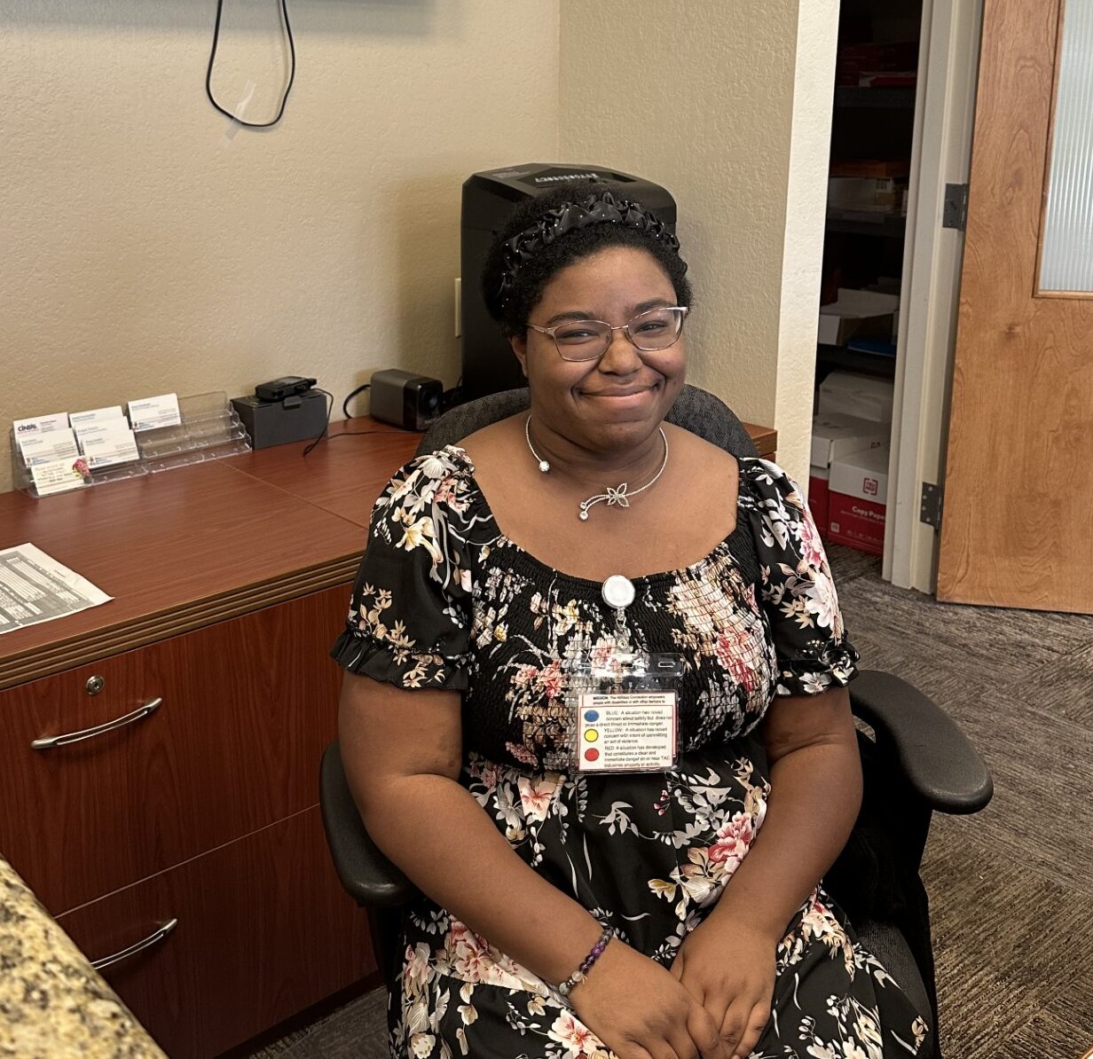 A person is sitting in an office setting, wearing a floral dress and a badge.
