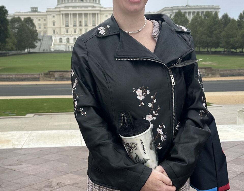 A person in a floral leather jacket holds a Starbucks cup outside a government building.