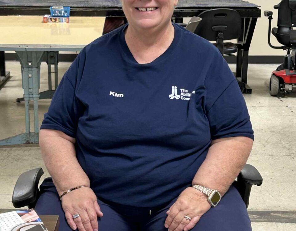 A person wearing a navy shirt with "Kim" and "The Ability Connection" sitting in a room with tables and chairs.