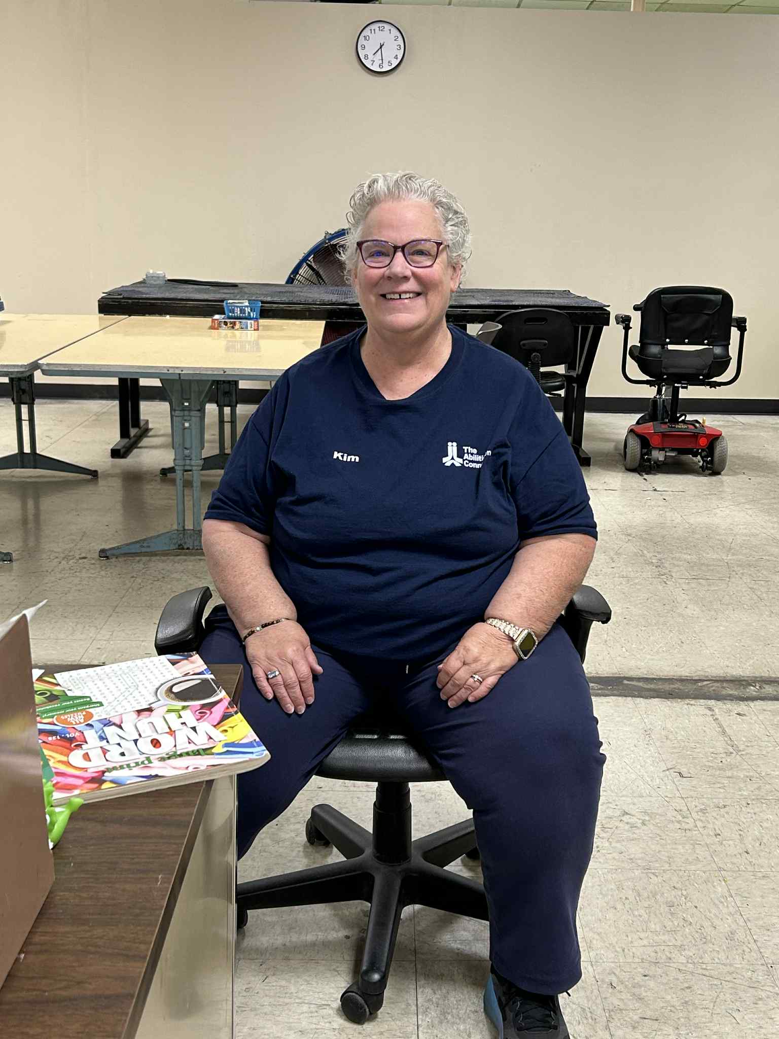 A person is sitting on an office chair in a room with tables, a clock, and a mobility scooter nearby.