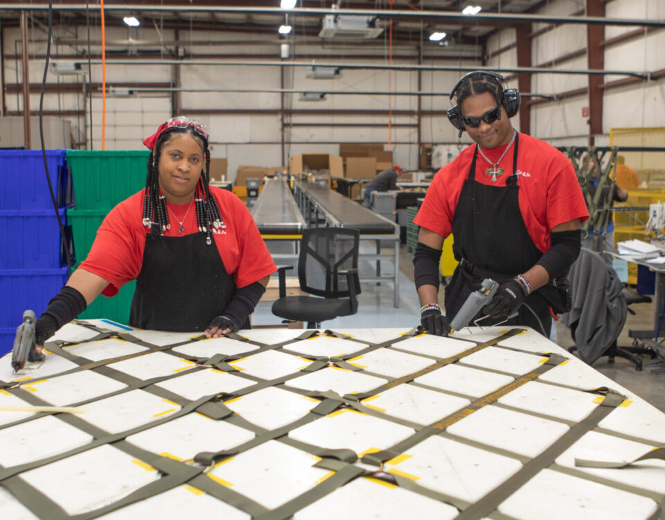 Two people in a workshop, wearing red shirts and black aprons, using tools on a strapped panel.