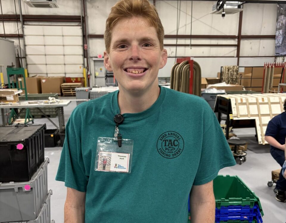 A person is smiling in a workshop or industrial setting, wearing a teal shirt and a badge.