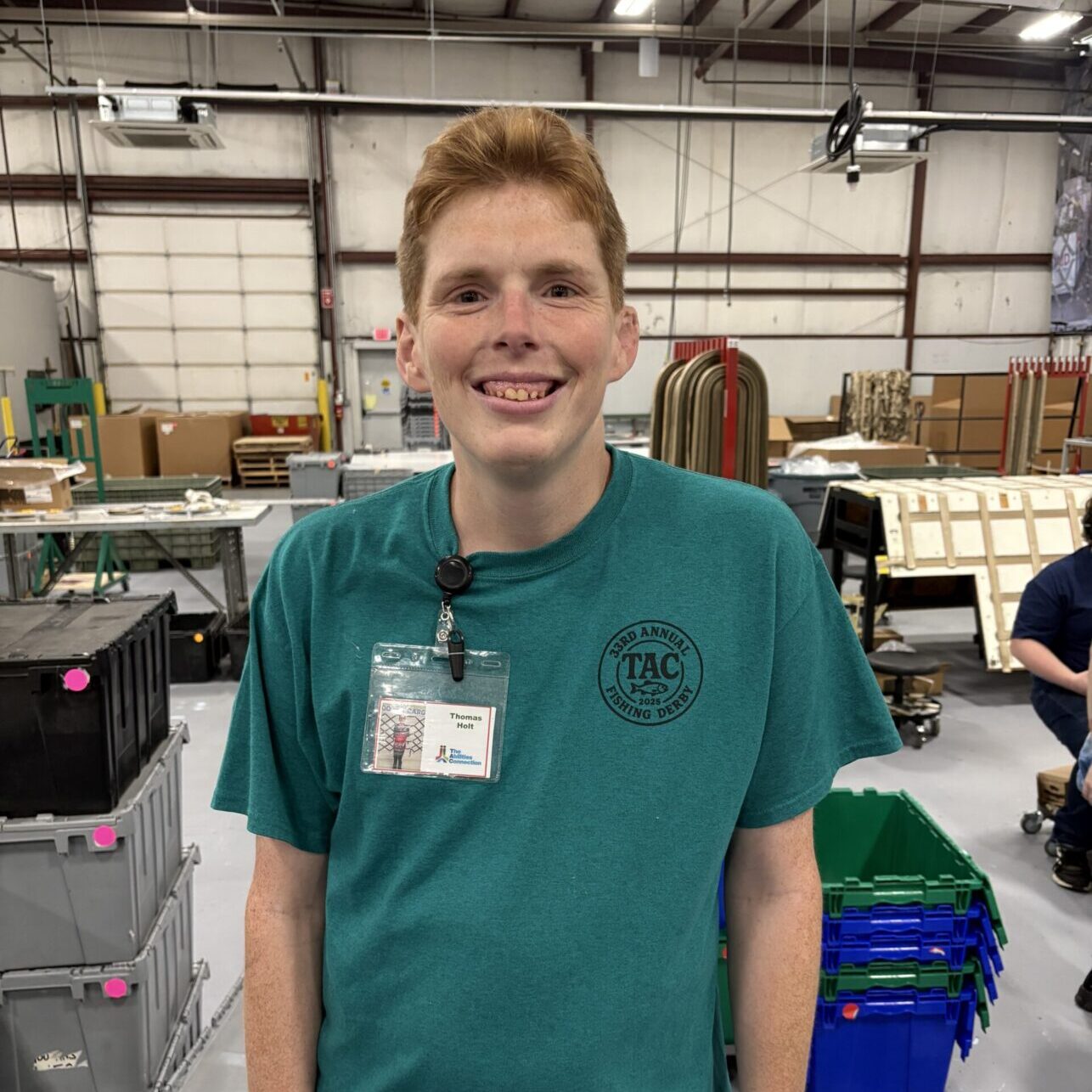 A person is smiling in a workshop or industrial setting, wearing a teal shirt and a badge.