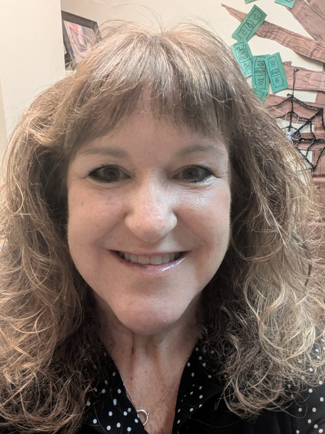 A person smiling with curly hair, wearing a necklace and a polka-dot shirt.