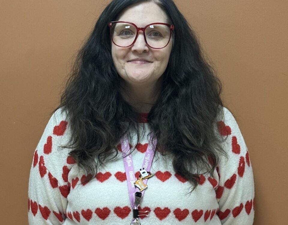 A person wearing glasses and a heart-patterned sweater, standing against a brown wall.