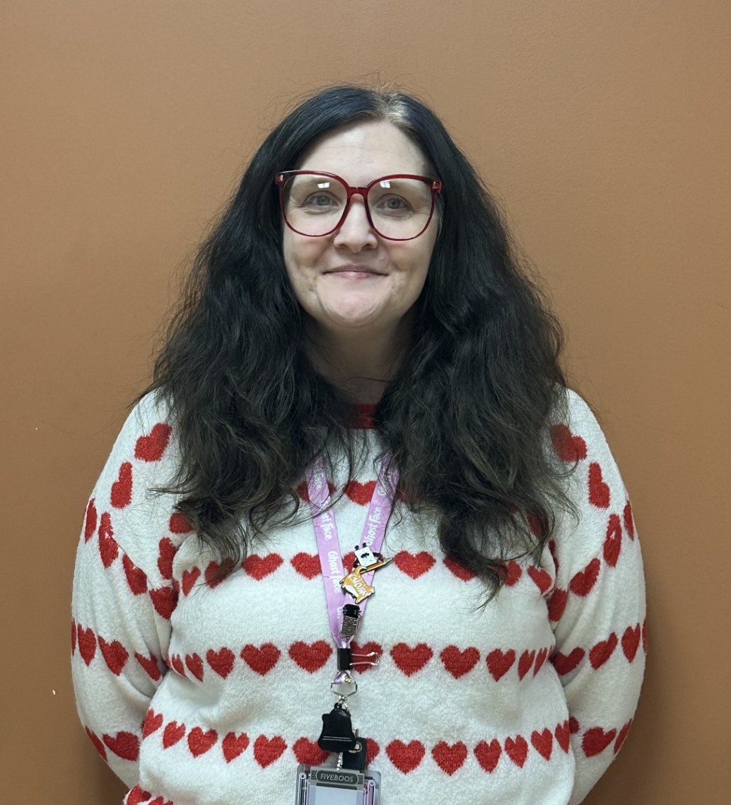 A person smiling with curly hair, wearing a necklace and a polka-dot shirt.