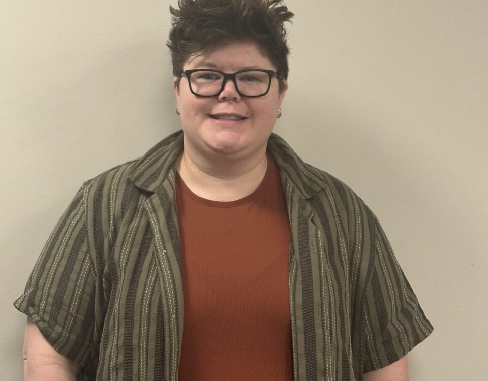A person with short hair, wearing glasses, a brown shirt, and a striped button-up.