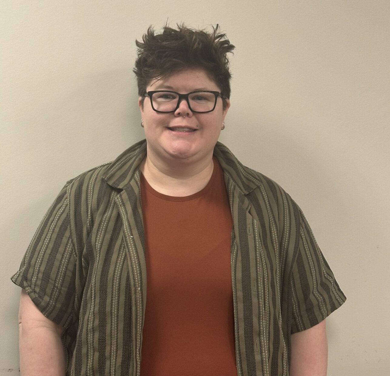 A person smiling with curly hair, wearing a necklace and a polka-dot shirt.