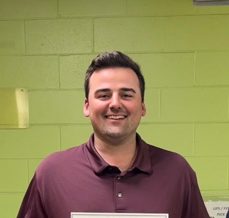 A person smiling, holding a certificate. The background features a green wall.