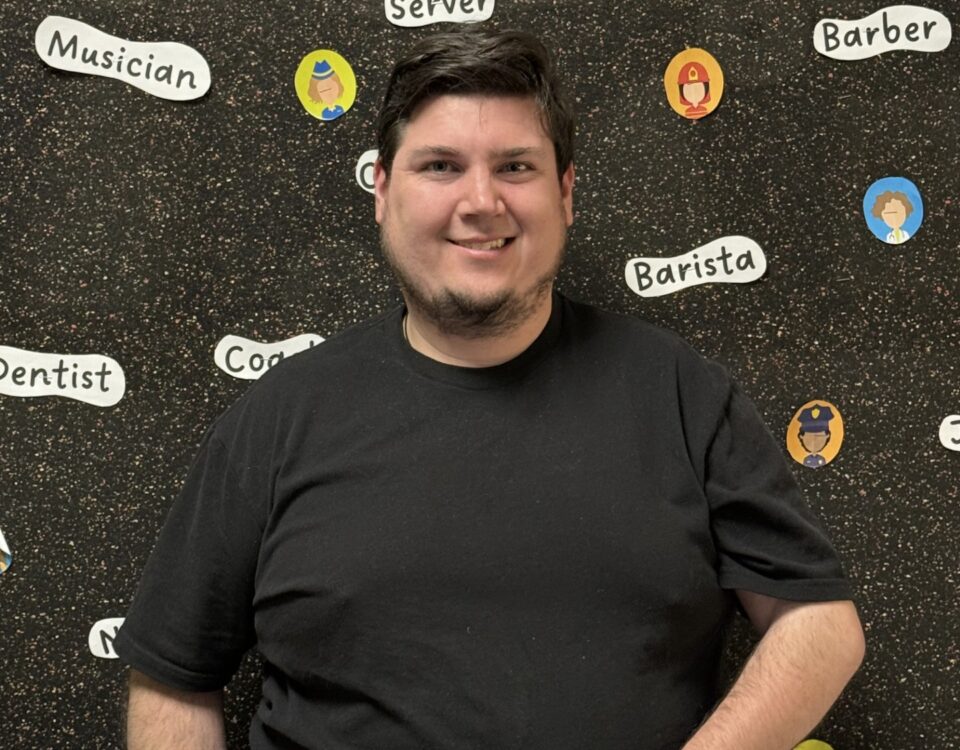 A person is smiling in front of a backdrop with various profession labels like "Musician," "Server," and "Barista."