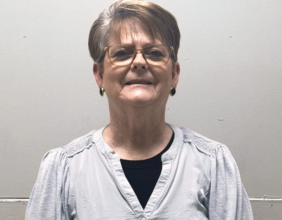 An older woman with glasses and short hair is smiling in front of a plain background.